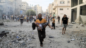 A Palestinian man carries a wounded girl at the site of Israeli strikes.png